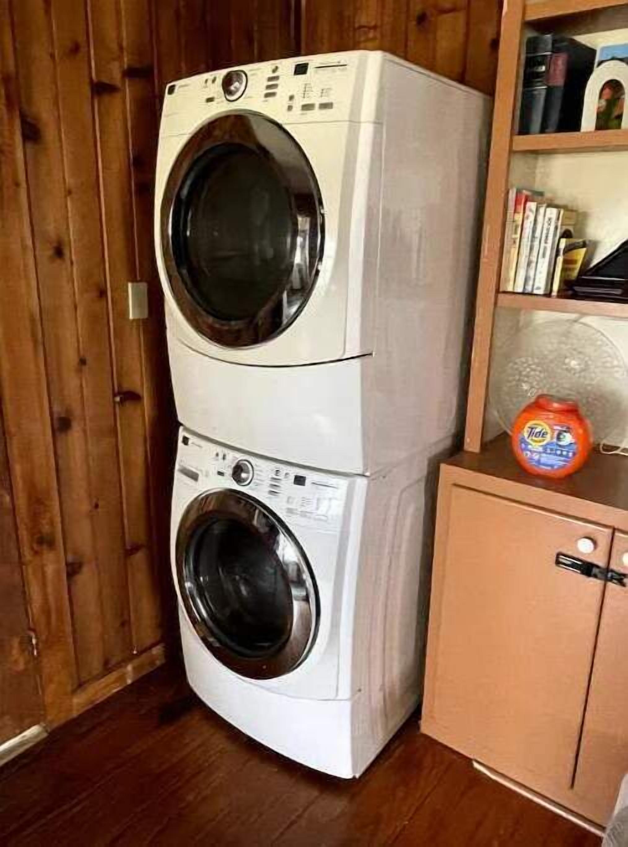 Une machine à laver et un sèche-linge superposés dans une pièce aux murs en bois, avec une petite armoire dotée d’une étagère.
