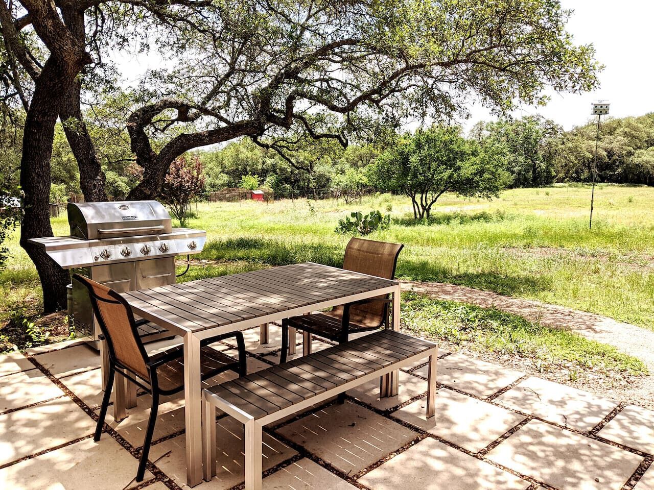 An outdoor patio with a wooden table and chairs, a barbecue grill, and a paved walkway.
