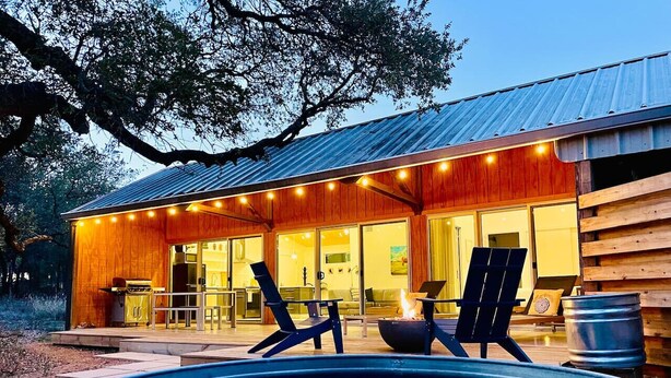 A pool with lounge chairs and a fire pit, surrounded by a wooden cabin with large windows and a metal roof. - Chic Barndo • Heated Pool, Firepit, Trails, Stars (Dripping Springs)
