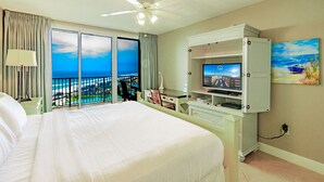 A hotel room with a large bed, a desk with a computer, a television, and a view of the ocean.