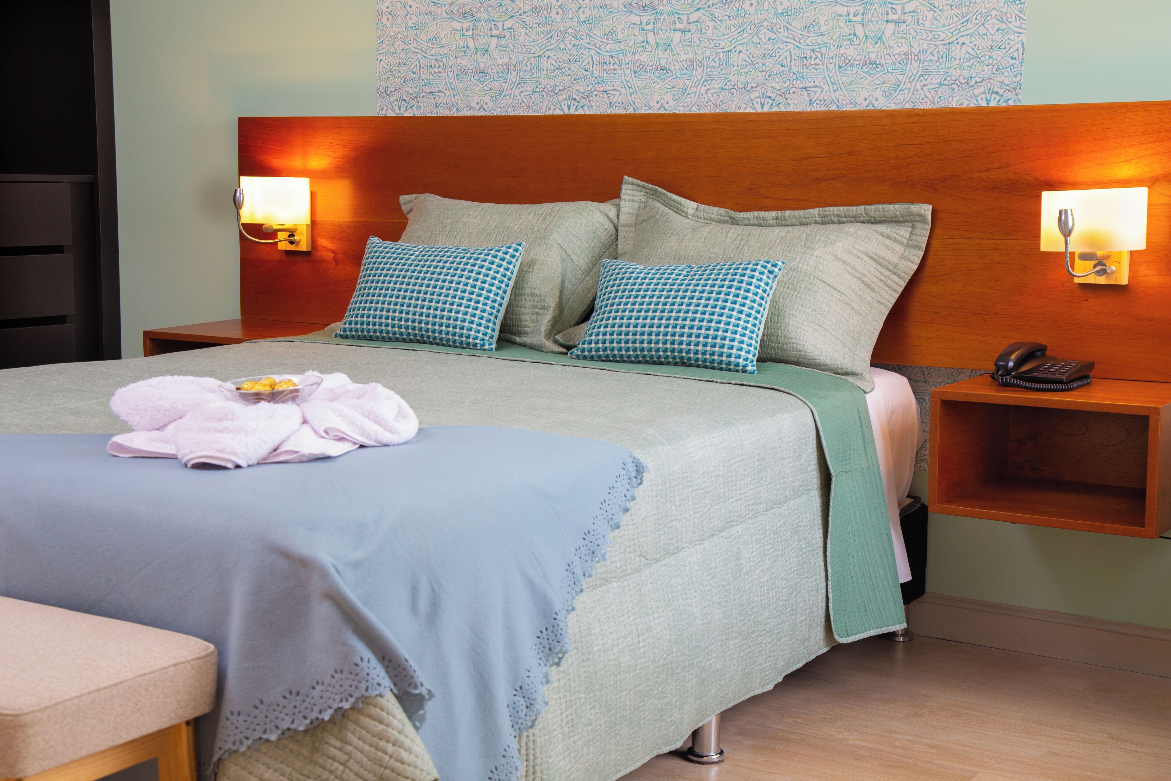 Cama bem arrumada com almofada azul e branca xadrez, manta verde e toalha rosa ao lado. Duas mesas de cabeceira, uma com telefone e outra vazia.