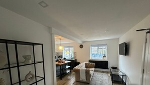 Uma sala de estar moderna com um sofá, uma mesa de centro e uma TV fixada na parede. Há uma área de jantar com mesa e cadeiras, e uma cozinha visível ao fundo.
