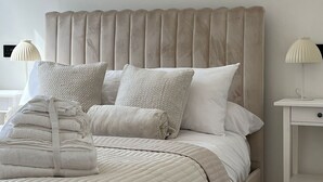 Cama bem arrumada com colcha acolchoada, travesseiros e toalhas dobradas. Um criado-mudo com uma luminária.