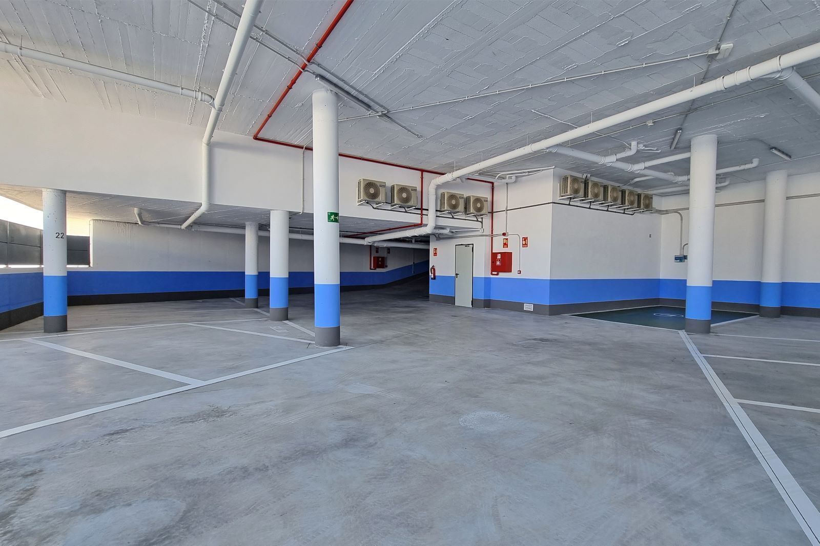 A spacious indoor parking area with concrete floors, blue and white walls, and multiple air conditioning units.