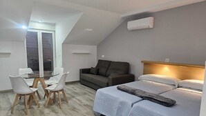 A modern, compact room with a bed, a sofa, a dining table with chairs, and a large window. - Apartamentos Antequera (Antequera)