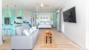 A modern living room with a sofa, a coffee table, and a TV. There is a kitchen area with a fridge, stove, and sink.