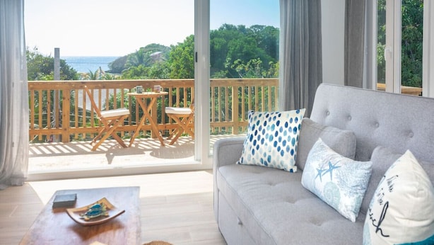 A balcony with wooden furniture, a grey sofa with patterned cushions, and a view of the sea and greenery.