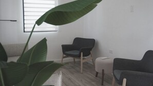 A living room with a large green plant, two dark chairs, a small ottoman, and a window with blinds. - Oceanfront Balcony King Bed suite steps to city center and beach Sunsets walks (Hatillo)