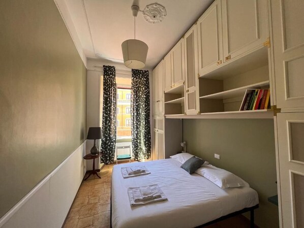 A bedroom with a bed, a nightstand, a lamp, a window with curtains, and built-in shelving with books.