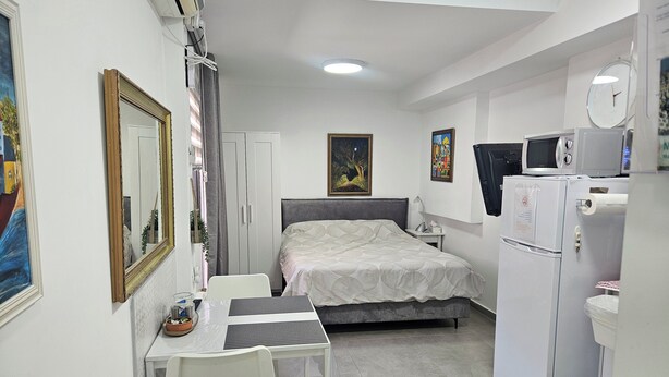 A bedroom with a bed, a desk, a chair, a wardrobe, a microwave, a refrigerator, and a trash bin. - Studio in Modi'in center (Modi'in-Maccabim-Re'ut)