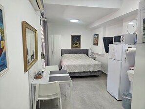 A bedroom with a bed, a desk, a chair, a wardrobe, a microwave, a refrigerator, and a trash bin. - Studio in Modi'in center (Modi'in-Maccabim-Re'ut)