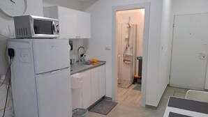 A compact kitchen with white cabinets, a microwave, and a refrigerator. A small trash bin and a mat are on the floor. - Studio in Modi'in center (Modi'in-Maccabim-Re'ut)