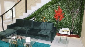 Un salon avec un canapé en velours vert, une table basse en verre et un escalier orné d’un mur vert et de fleurs rouges.