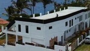 Un edificio blanco con múltiples ventanas y techo plano, rodeado de palmeras y vista al océano.