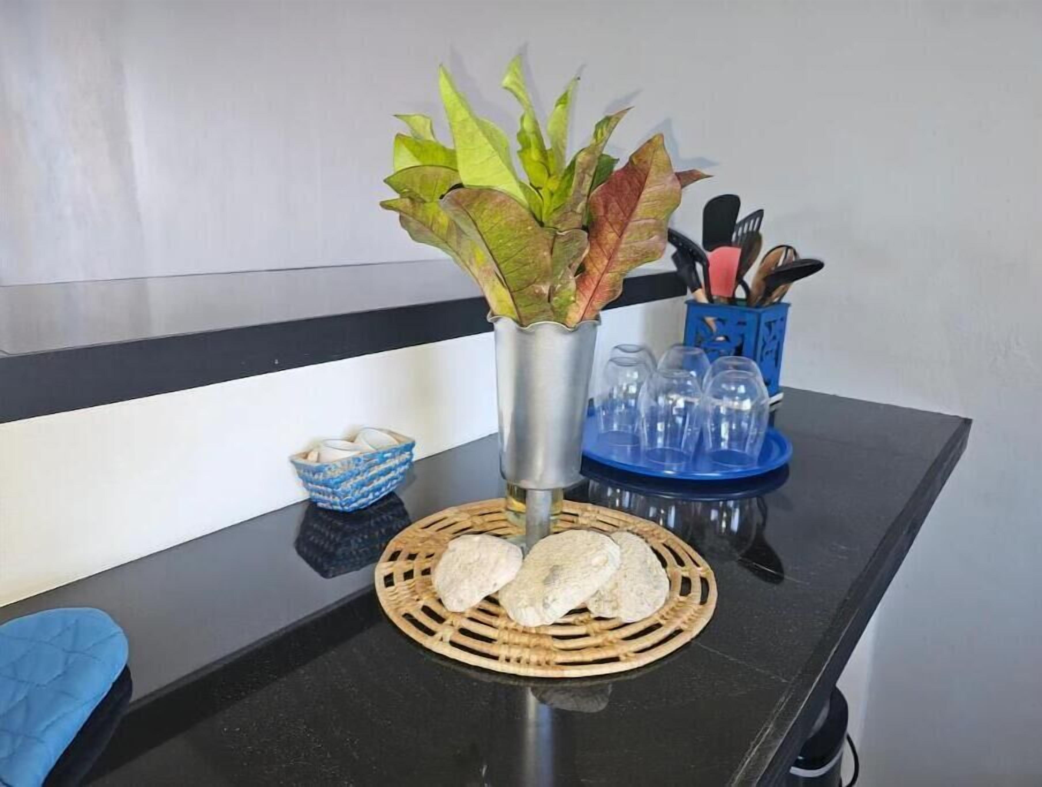 Una encimera negra con un mantel tejido que sostiene dos tortillas, un tazón azul pequeño, un florero de metal con hojas verdes y un recipiente azul que contiene utensilios.
