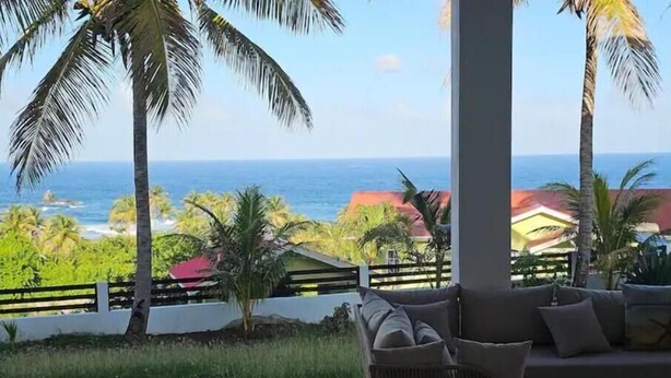 Un patio con un sofá de mimbre y una mesa redonda, con vistas al mar, palmeras y casas.