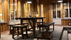 Une terrasse extérieure en bois, dotée de tables de pique-nique et de bancs, d’une lanterne, et offrant une vue sur l’intérieur grâce à de grandes fenêtres.