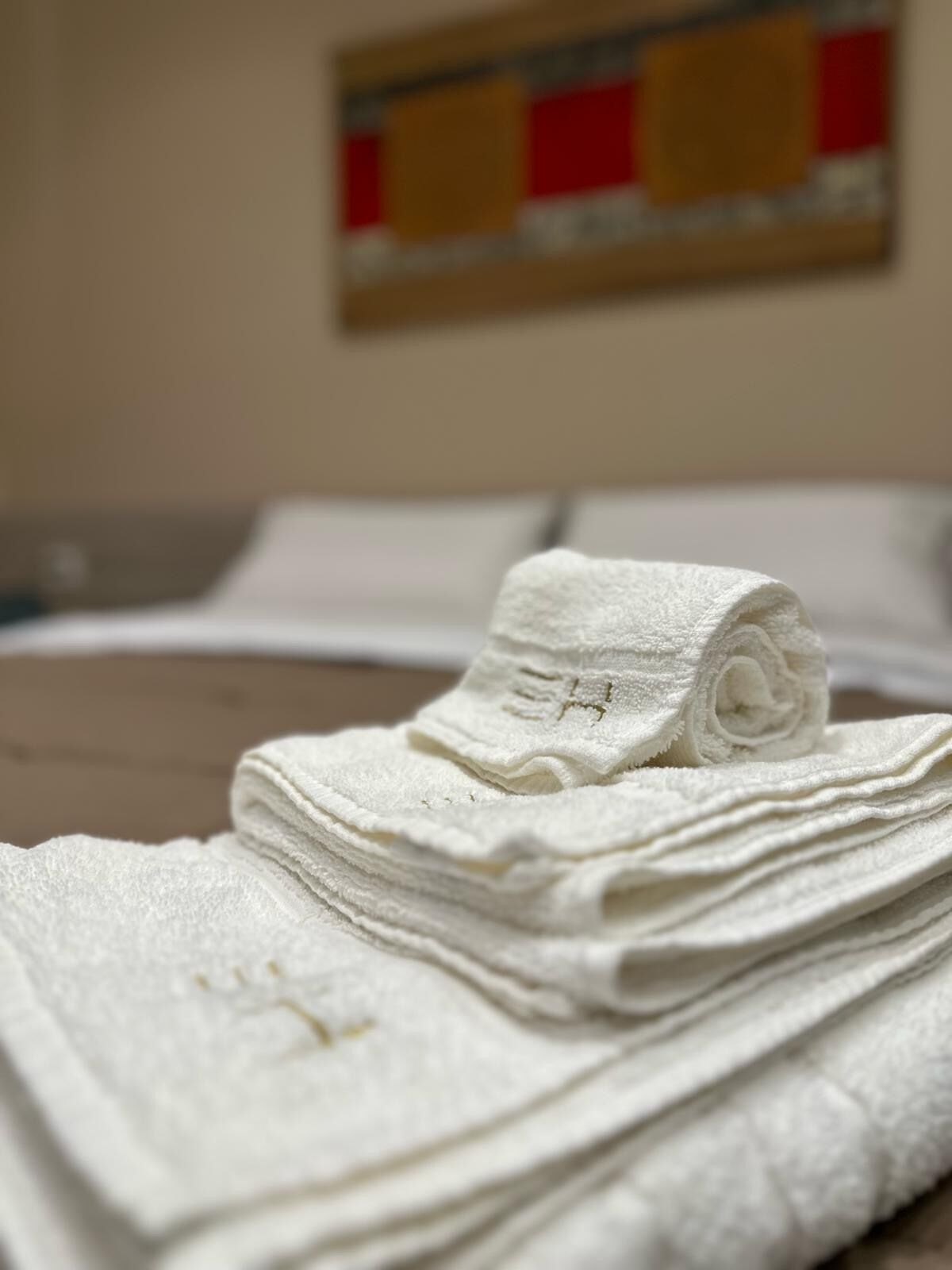 A neatly folded white towel on a bed with a plain wall and a painting in the background.