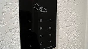Un panel de teclado electrónico negro con un indicador luminoso rojo, montado sobre una pared blanca texturizada.