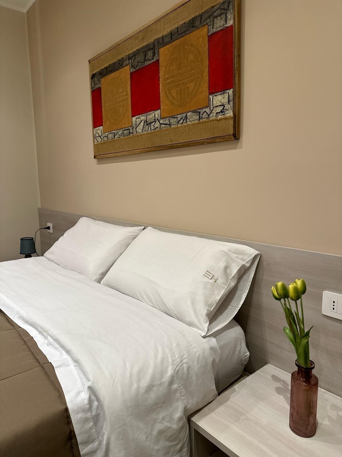 Ein Hotelzimmer mit Bett, Nachttisch, Blumen in einer Vase und einem Wandbild.