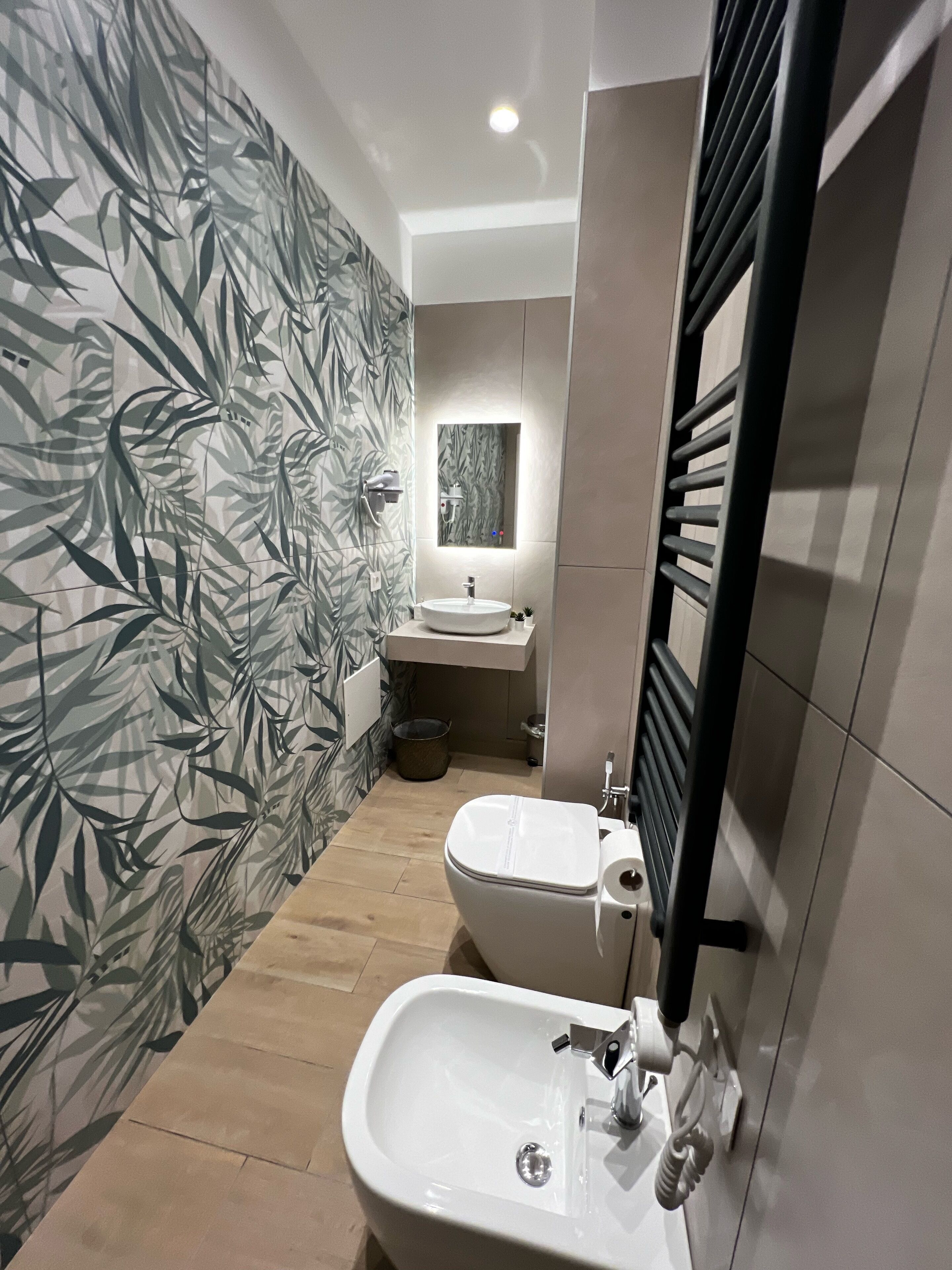 A modern bathroom with a toilet, bidet, and sink. The walls have a bamboo leaf wallpaper, and there is a large mirror above the sink.
