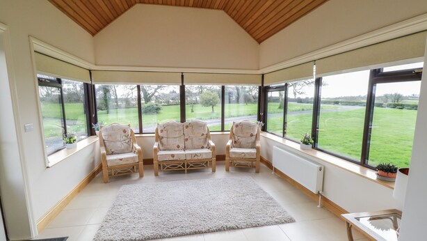 A sunroom with a high ceiling, large windows, and three wicker chairs. - Bunny Lane (Cloonflyn)