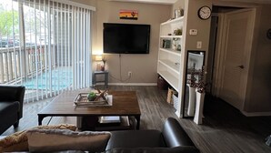 A living room with a flat-screen TV mounted on the wall, a coffee table with decorative items, a sofa, and a sliding glass door leading to a balcony.