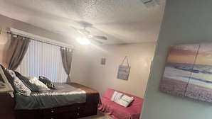 A bedroom with a bed, a couch, and a small table. There is a ceiling fan, a window with curtains, and a wall-mounted air conditioning unit.