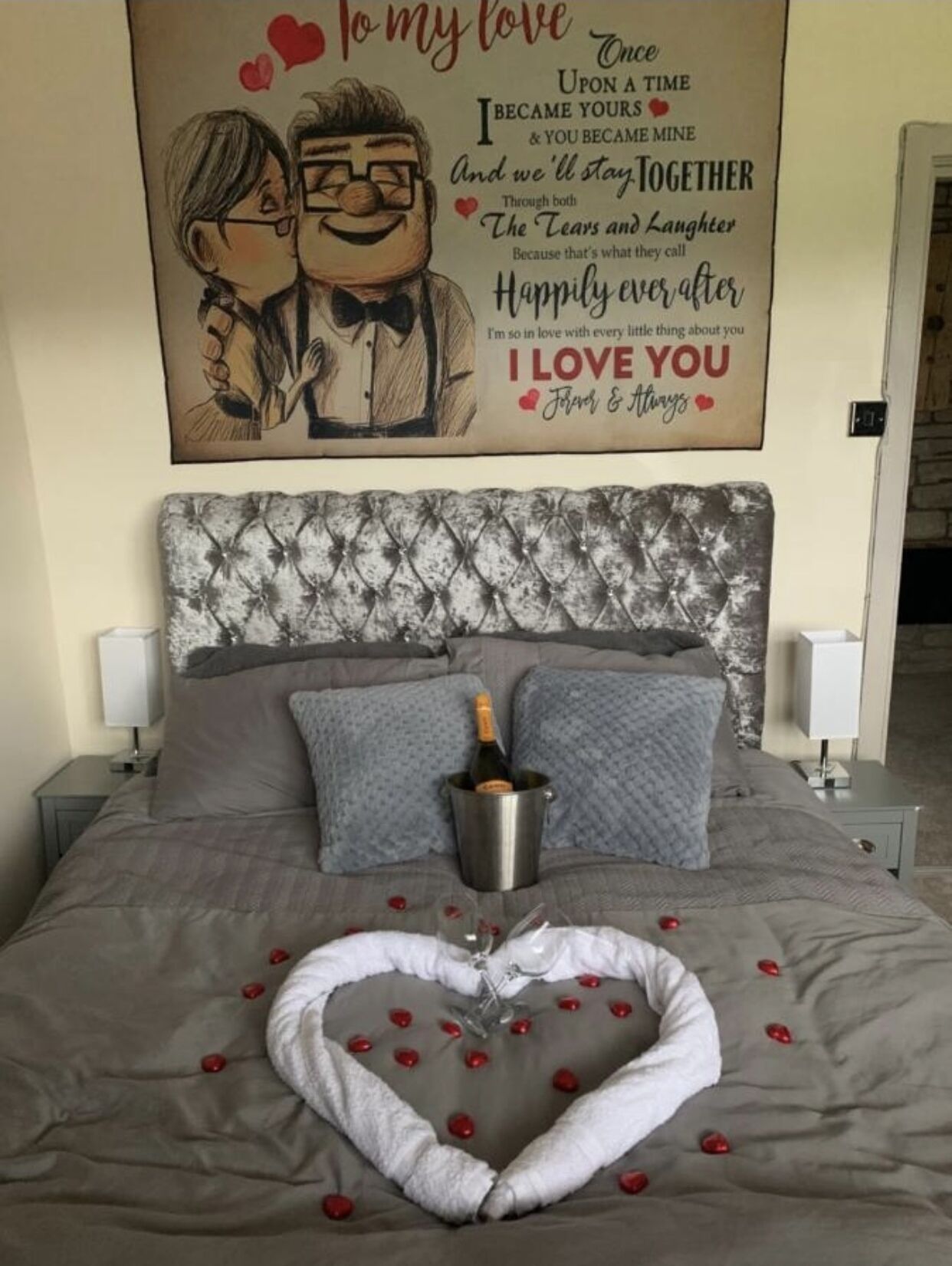 A neatly made bed with a heart-shaped arrangement of rose petals and a bottle of champagne in a bucket.