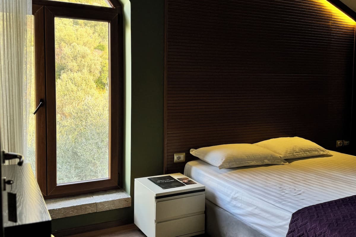 Un dormitorio con un ventanal grande, una cama con ropa de cama blanca, una mesita de noche, una pequeña mesa y un tapete estampado.