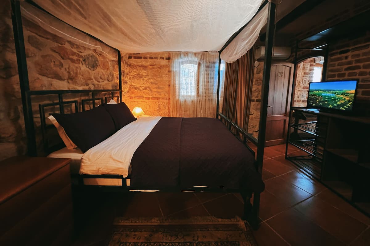 Un dormitorio con cama con dosel, paredes de piedra, televisor y ventana con cortinas.