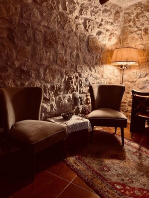 A cozy room with a stone wall, two armchairs, a small table with a bowl, and a lamp.