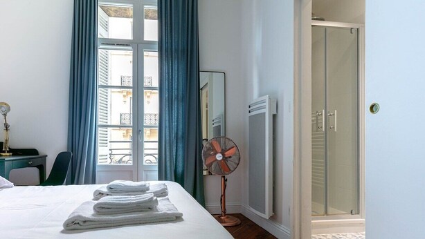 Een hotelkamer met een bed, een ventilator, een raam met gordijnen, een spiegel, een radiator en een douche.
