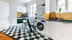 Een kleine keuken met een wasmachine, een eethoek met een geruit vloertje en een raam met een plant.