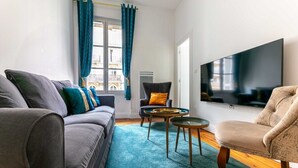 Een moderne woonkamer met een grijze bank, een beige fauteuil en een zwarte fauteuil met een gouden kussen. Een grote flatscreen televisie is aan de muur bevestigd.