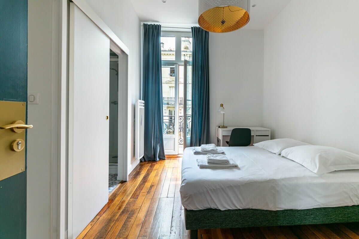 A bedroom with a wooden floor, a bed with white linens, a desk, and a chair.