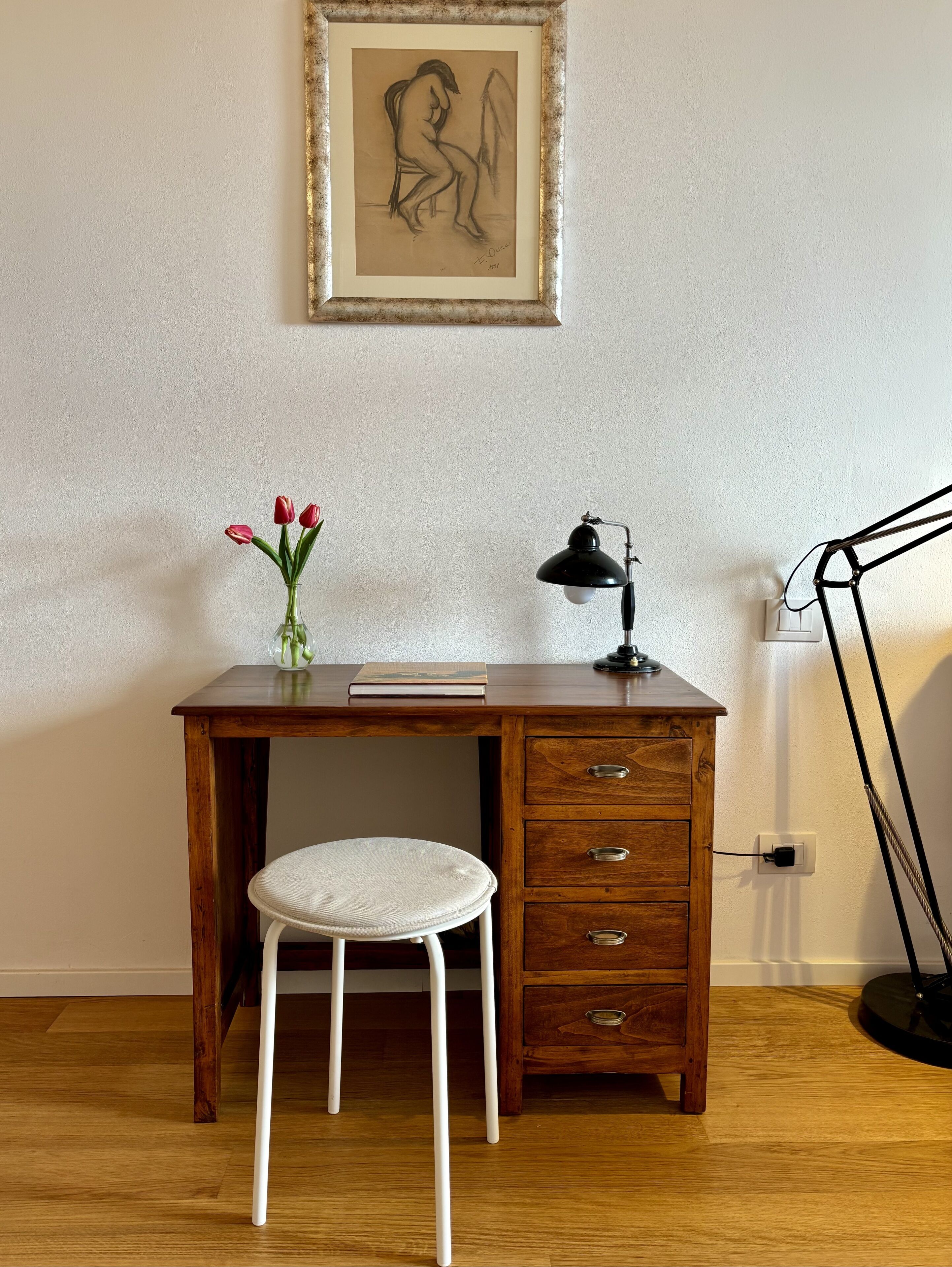 Un bureau en bois, accompagné d’un tabouret blanc, d’une lampe et d’un vase contenant des fleurs rouges.