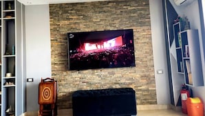 A living room with a flat-screen TV mounted on a stone wall, a wooden chair, a dark sofa, and a bookshelf. - Pyramids planet hotel (Giza)