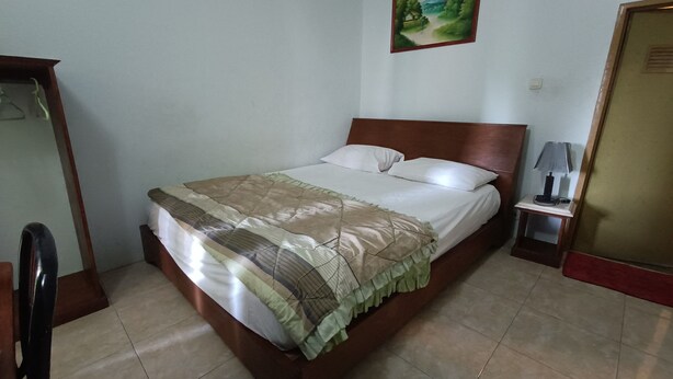 A neatly made bed with a patterned bedspread and pillows, a small bedside table with a lamp, and a painting on the wall. - Hotel O Vj Sweethome Syariah (Mataram)