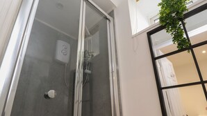 A glass-enclosed shower with a handheld showerhead, a white wall, and a window with a black frame. - Basement Flat 43 (Keighley)