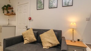 A grey sofa with two beige cushions, a bedside table with a lamp, and three framed botanical prints on the wall. - Basement Flat 43 (Keighley)