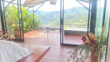 Una habitación con suelo de madera, una cama, un sillón de mimbre y vistas a montañas a través de amplios ventanales.