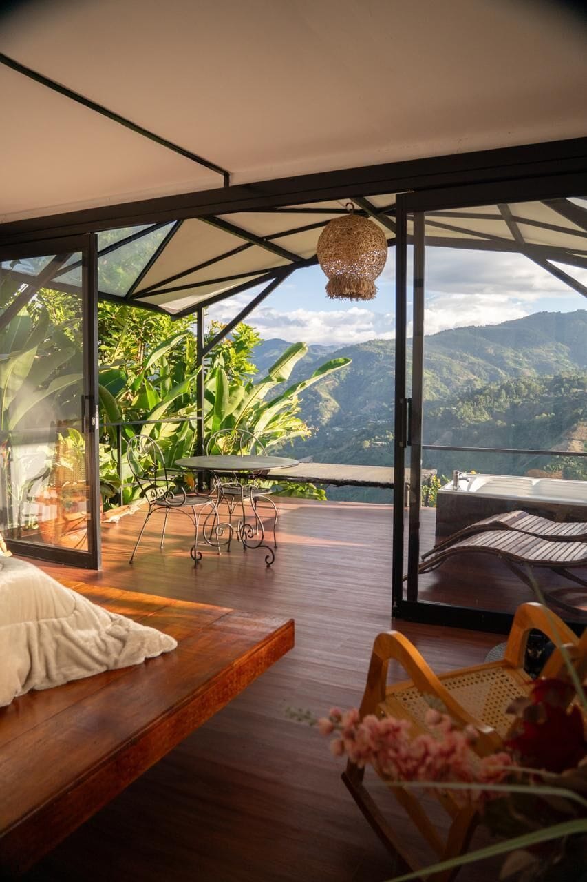 Habitación con piso de madera, una cama, una mesa y sillas. Se observa una vista de montañas y vegetación desde el exterior.