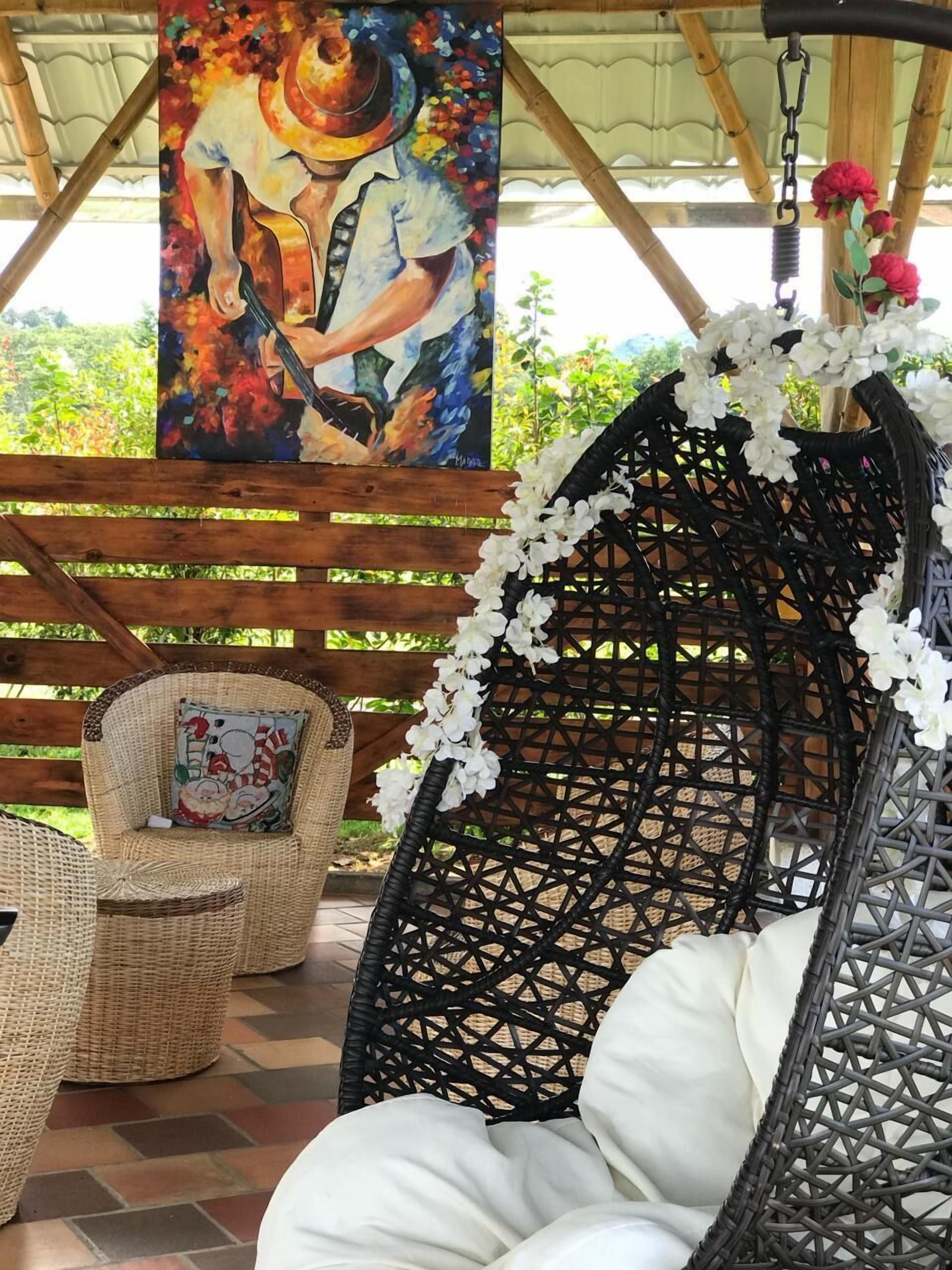 A wicker chair with white cushions and a hanging flower arrangement, a wicker sofa with a colorful cushion, and a painting of a person playing a guitar.