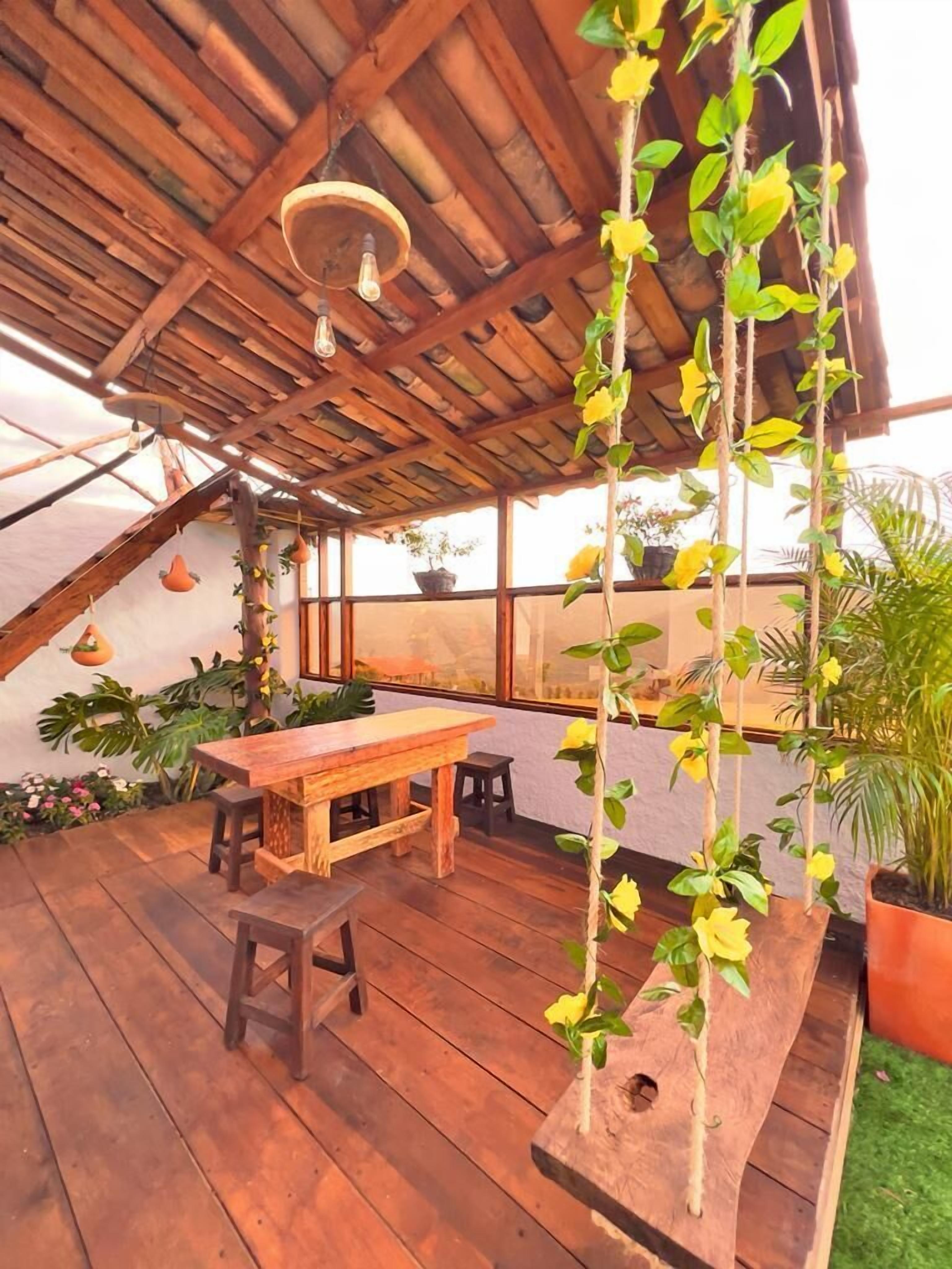 Une terrasse extérieure en bois, avec une table et des bancs en bois, entourée de plantes en pot et de plantes grimpantes suspendues.