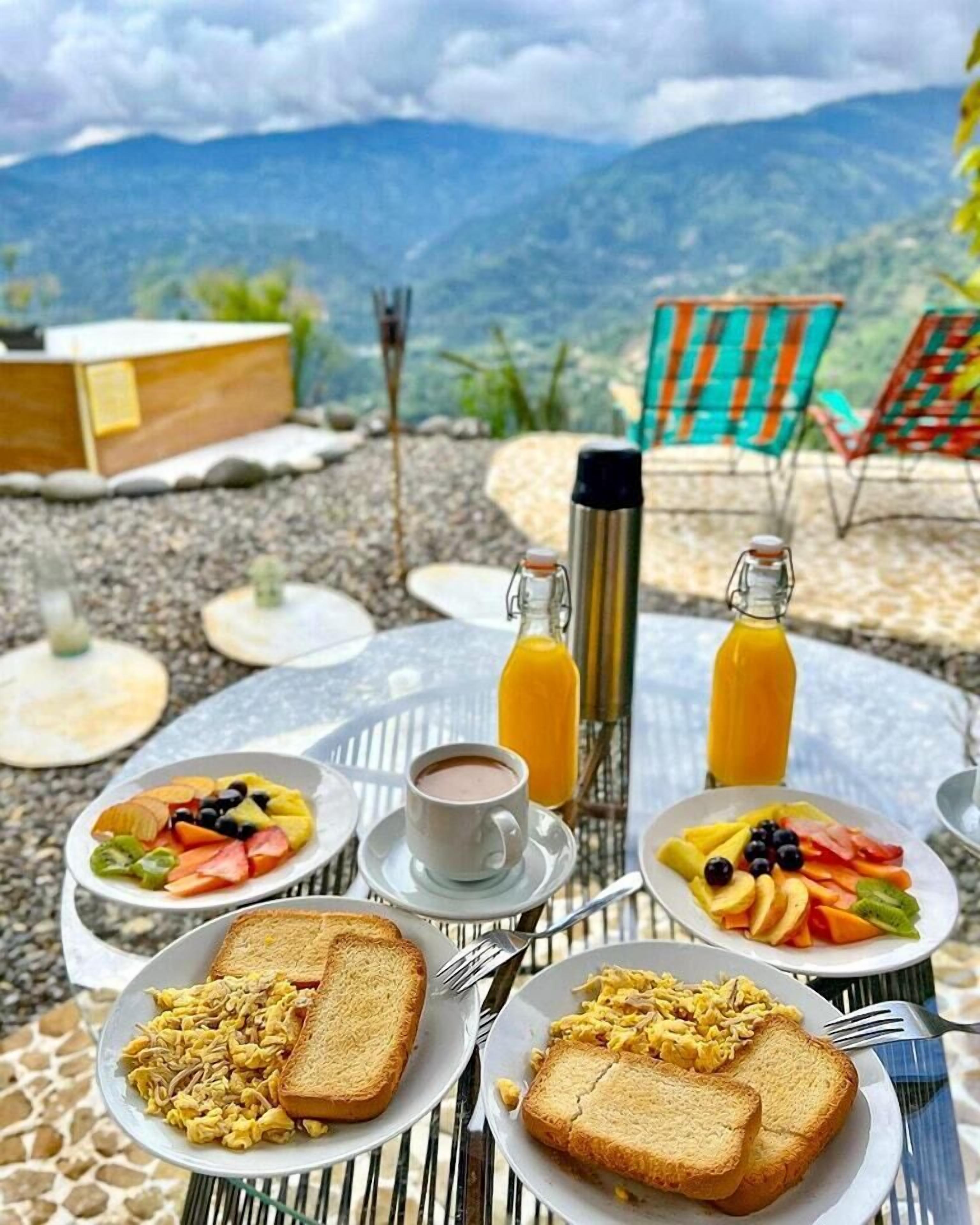 Ein Frühstückstisch im Freien mit Toast, Rührei, Obst, einer Tasse Kaffee und zwei Orangensaftflaschen, mit Blick auf eine Berglandschaft.