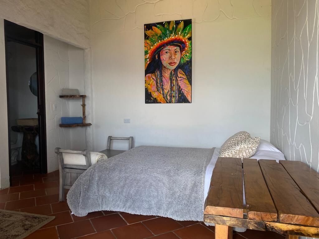 A bedroom with a bed, a wooden table, a chair, a shelf, and a painting on the wall.
