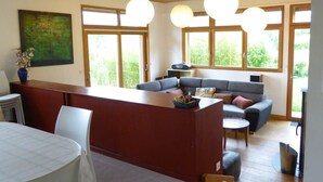 A modern living room with a wooden ceiling, large windows, a grey sofa, a dining table, and a kitchen area. - Chalet avec grand jardin en Chartreuse (Alpes) (Miribel-les-Échelles)