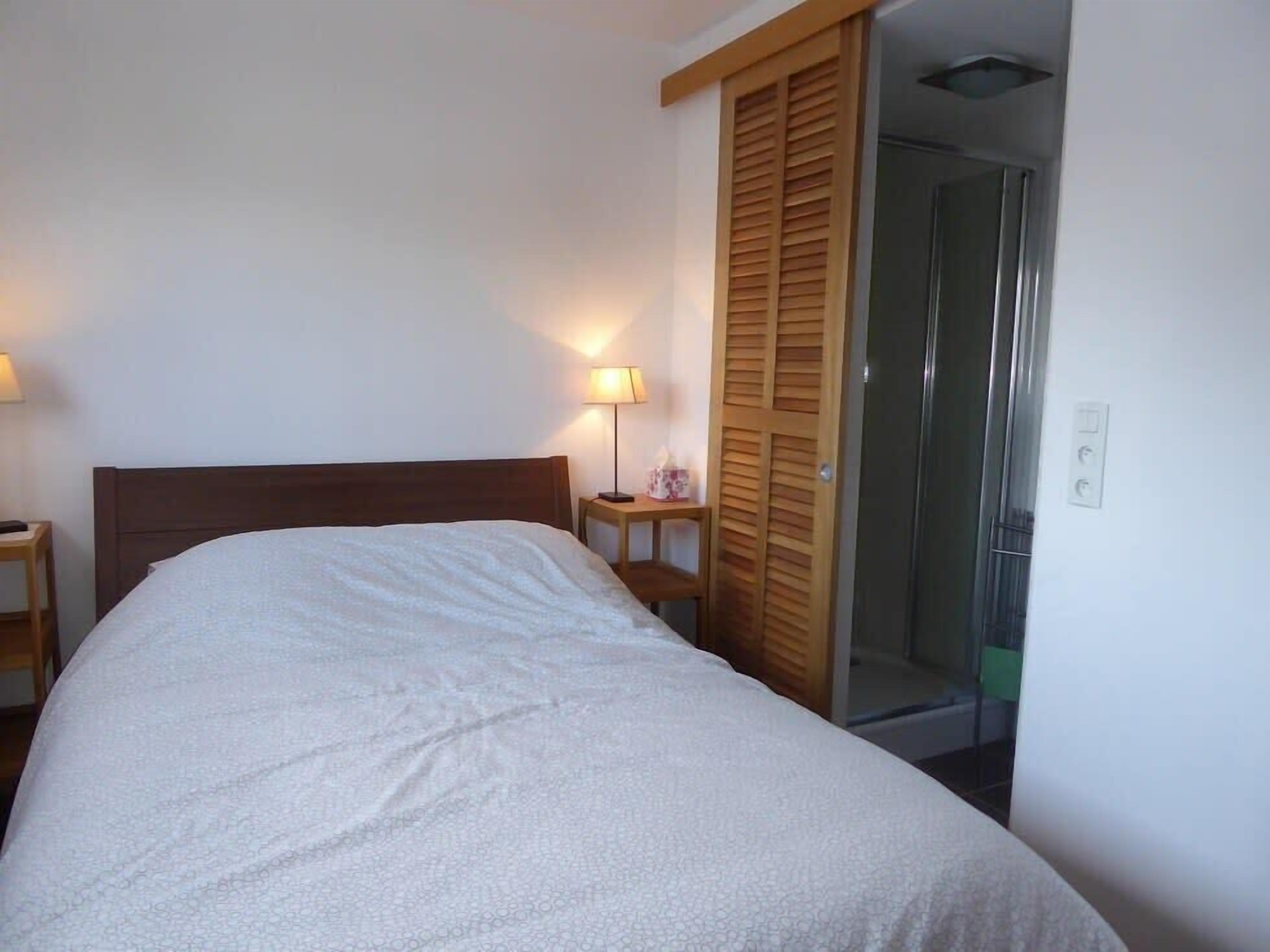 Une chambre à coucher comprenant un lit, des lampes de chevet, une armoire en bois et une porte en verre donnant sur un espace douche.