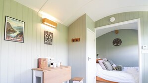 Ein gemütliches Schlafzimmer mit einem Bett, einem Schreibtisch mit Stuhl und einem kleinen Hocker. An der Wand hängt ein gerahmtes Bild und ein Schild mit der Aufschrift 'All you need is love & good coffee.'